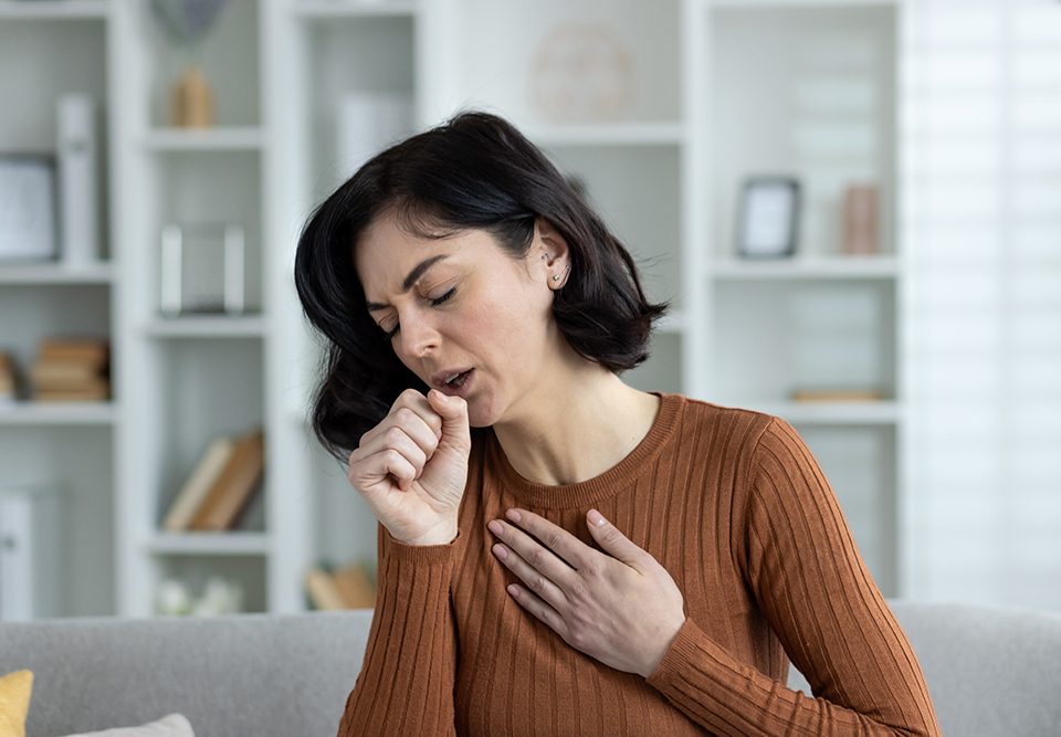 Foto em close-up de uma jovem em casa, sentada no sofá e tossindo forte, sentindo dor no peito