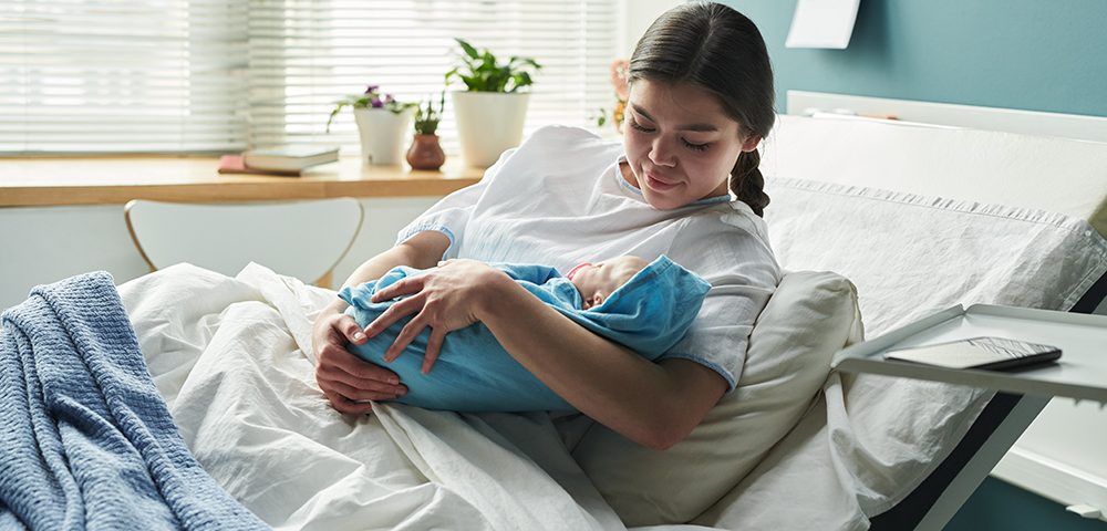 Jovem mãe sentada na cama de hospital segurando bebê recém-nascido